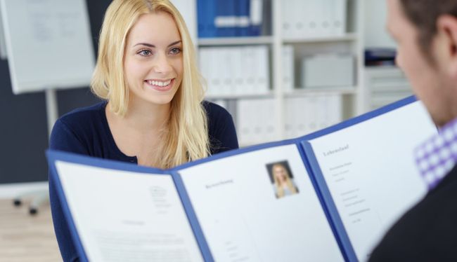 Attractive young businesswoman in a job interview with a corporate personnel manager who is reading her CV in a blue folder, over the shoulder focus to the young applicant